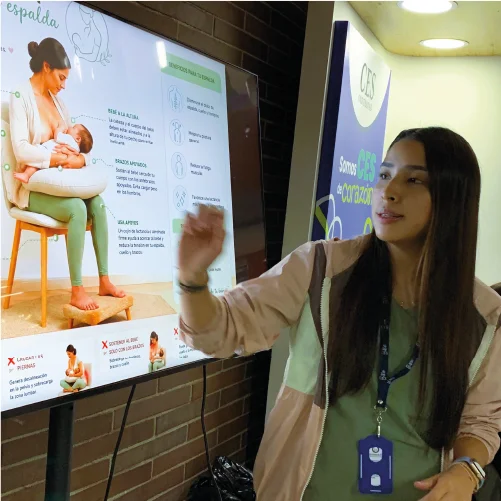 Persona realiza una presentación junto a una pantalla digital que muestra información ilustrada sobre lactancia materna, en un espacio institucional con material educativo visible.