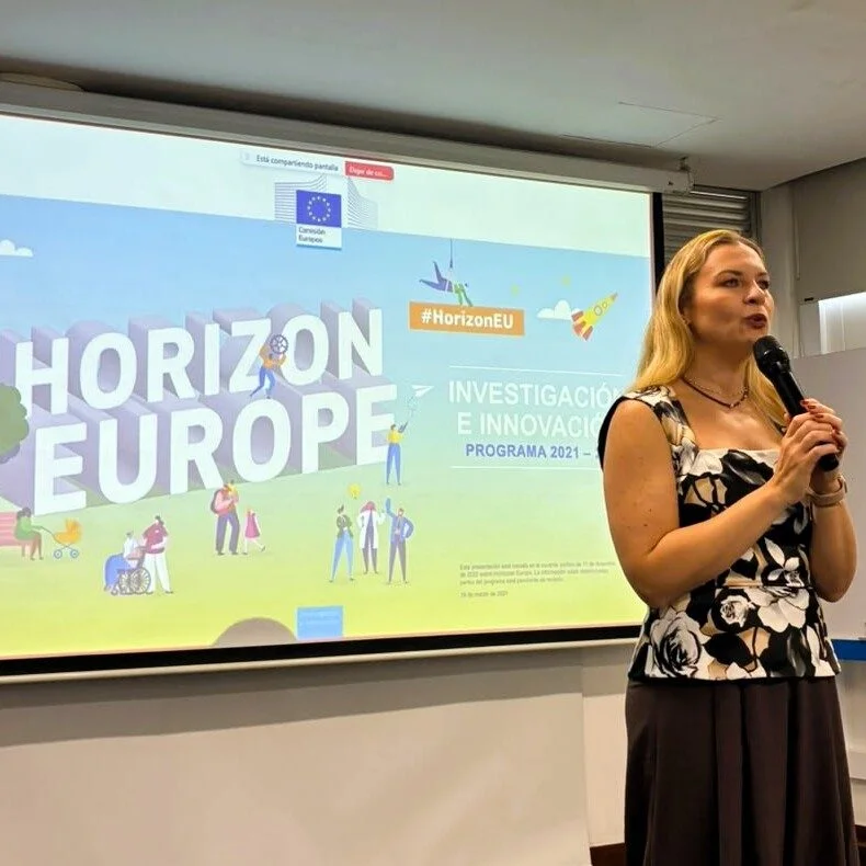 Persona realizando una presentación en un aula o sala de conferencias, frente a una pantalla proyectada con información sobre el programa europeo de investigación e innovación Horizon Europe. A un lado se observa una pizarra blanca. La imagen ilustra una actividad académica o informativa relacionada con oportunidades de investigación y cooperación internacional.