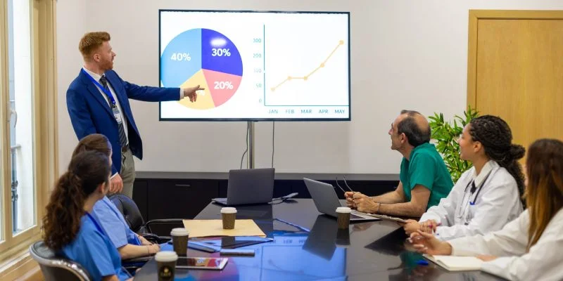 Grupo de personas reunidas alrededor de una mesa de conferencias mientras una persona presenta gráficos estadísticos en una pantalla. La escena muestra una reunión de análisis de datos en un entorno profesional del sector salud.