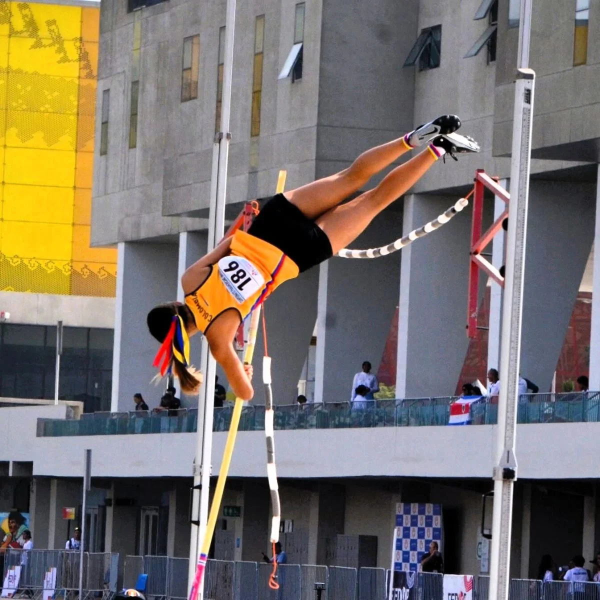 Persona atleta realiza un salto con pértiga durante una competencia deportiva al aire libre. La imagen muestra el momento en que el cuerpo supera el listón horizontal, con instalaciones deportivas y un edificio moderno al fondo.