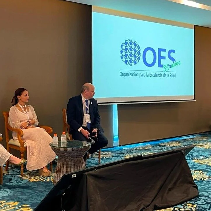 Panel académico o institucional con dos personas sentadas frente a una pantalla de proyección que muestra el logo de la Organización para la Excelencia de la Salud (OES) y la conmemoración de sus 35 años. La actividad se desarrolla en un salón de conferencias con mobiliario para exposiciones y diálogo entre expertos.