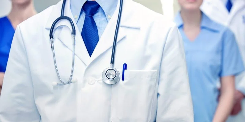Primer plano de profesionales de la salud en un entorno hospitalario. En el centro se observa una bata médica blanca con un estetoscopio alrededor del cuello, mientras que al fondo aparecen otras personas con indumentaria clínica.