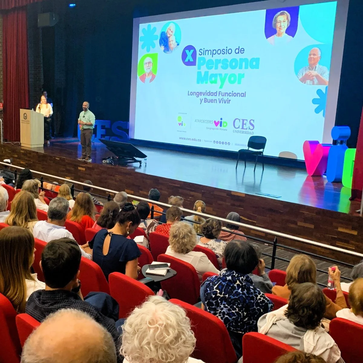 Fotografía de un auditorio durante un evento académico sobre personas mayores. En el escenario, una persona realiza una presentación frente a una pantalla que muestra el título “X Simposio de Persona Mayor: Longevidad Funcional y Buen Vivir”, junto con los logotipos de la Universidad CES y entidades aliadas. El público ocupa gran parte de las sillas rojas del auditorio, observando la exposición.