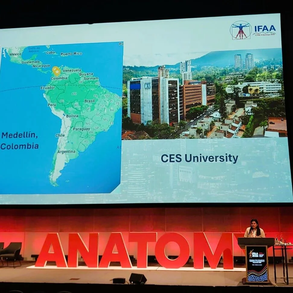Diapositiva de una presentación sobre la Universidad CES en Medellín, Colombia. A la izquierda aparece un mapa de América Latina con la ubicación de Medellín señalada en Colombia. A la derecha se muestra una fotografía aérea del campus universitario. En la parte inferior se lee “CES University”. La diapositiva se proyecta en un auditorio durante un evento de anatomía, con un ponente en un atril y grandes letras rojas que forman la palabra “ANATOMY” frente al escenario.