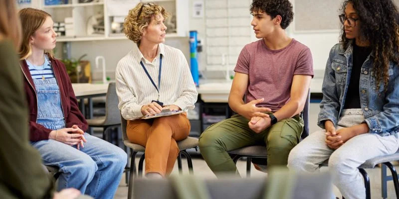 Grupo de estudiantes y una persona facilitadora sentados en círculo dentro de un aula, participando en una conversación. El espacio contiene mobiliario escolar y estanterías con materiales al fondo.