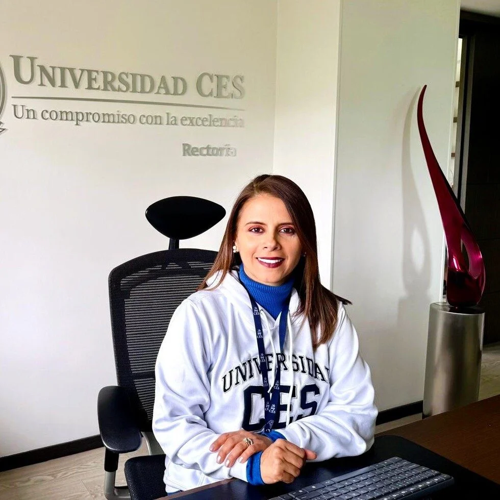 Persona sentada en un escritorio dentro de una oficina institucional de la Universidad CES. En la pared se observa el texto “Universidad CES” y “Rectoría”. La persona viste una sudadera blanca con el logo de la universidad y porta una escarapela. Sobre el escritorio hay un teclado y, al fondo, una escultura decorativa de color rojo.