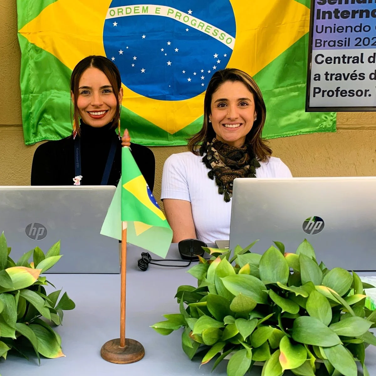 Dos personas están sentadas detrás de una mesa con computadores portátiles. Al fondo se observa una bandera de Brasil y, en primer plano, una pequeña bandera del mismo país junto a plantas decorativas. La escena corresponde a un espacio informativo o académico relacionado con Brasil.