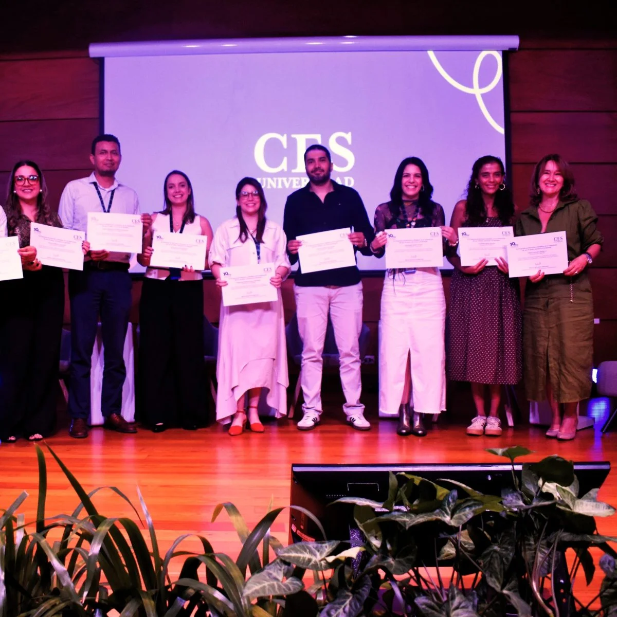 Grupo de personas sobre un escenario sosteniendo certificados durante una ceremonia académica en la Universidad CES, frente a una pantalla con el logo institucional.