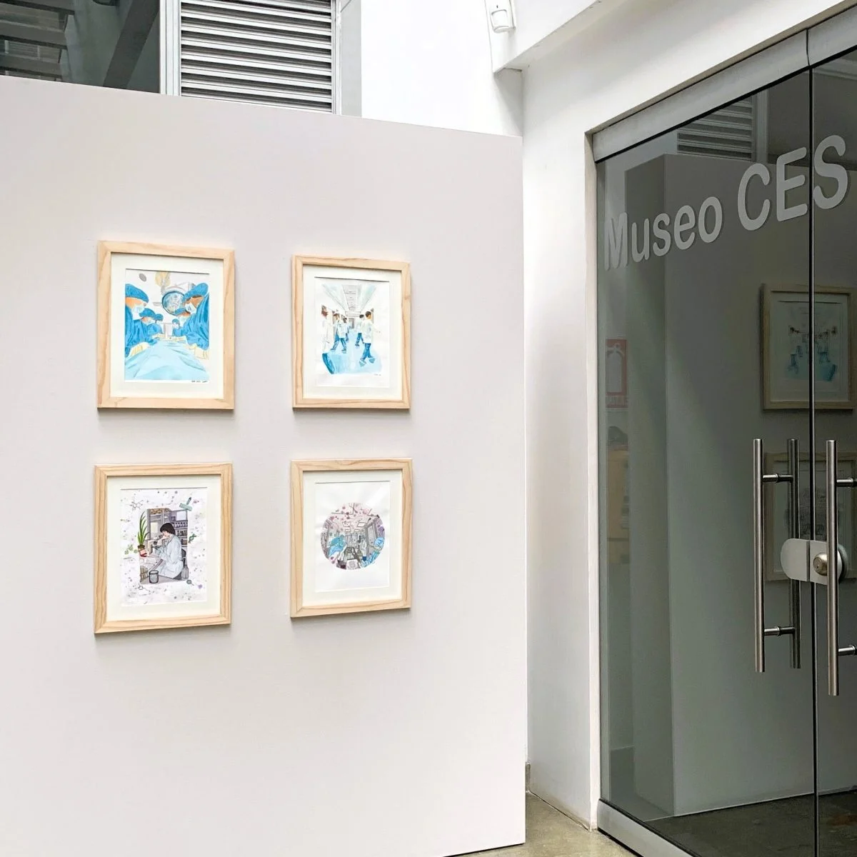 Vista de la entrada del Museo CES con una muestra de cuatro ilustraciones enmarcadas exhibidas sobre un panel blanco junto a una puerta de vidrio.
