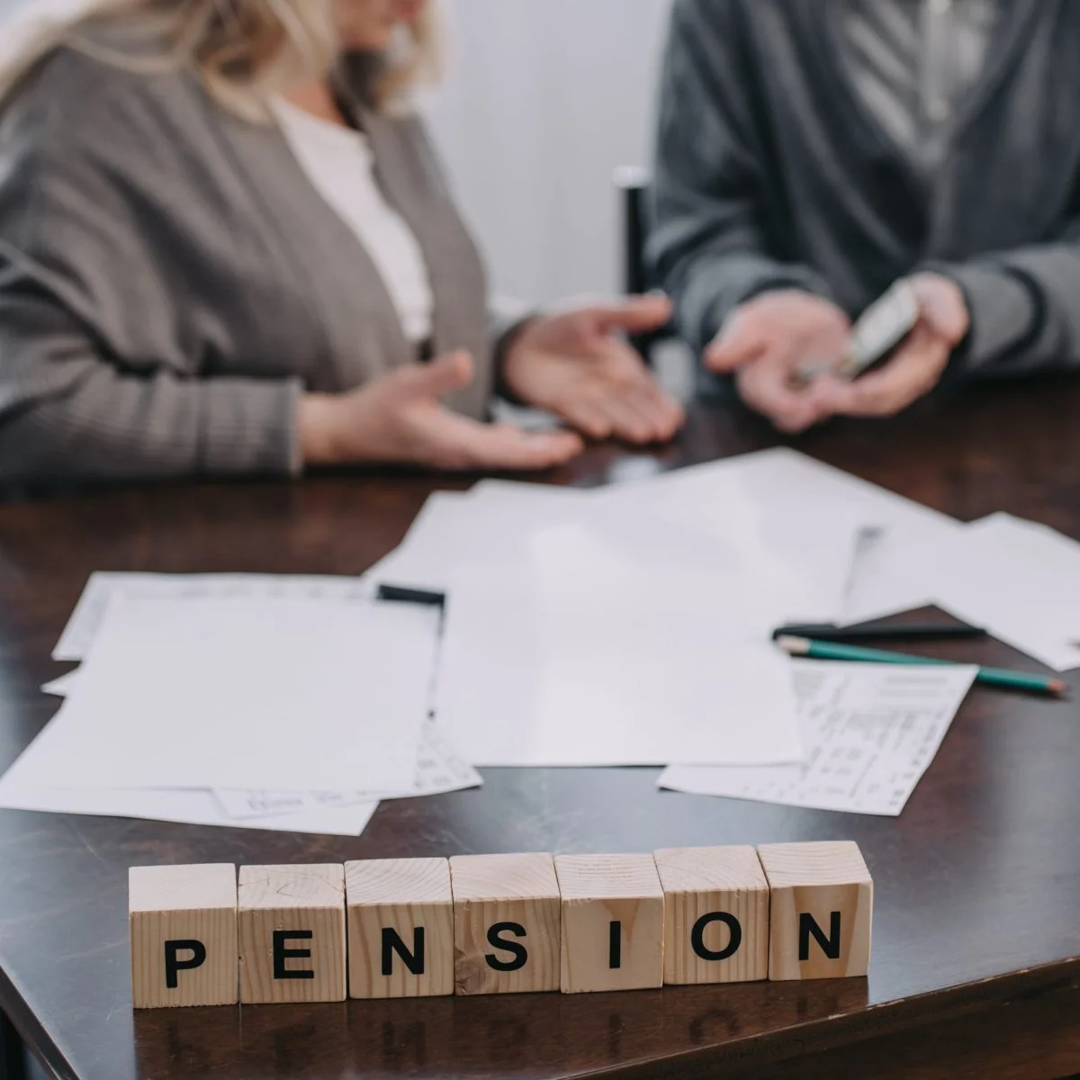 Dos personas conversan en una mesa de madera con documentos, bolígrafos y un teléfono móvil. En primer plano, unos bloques de madera forman la palabra “PENSION”.