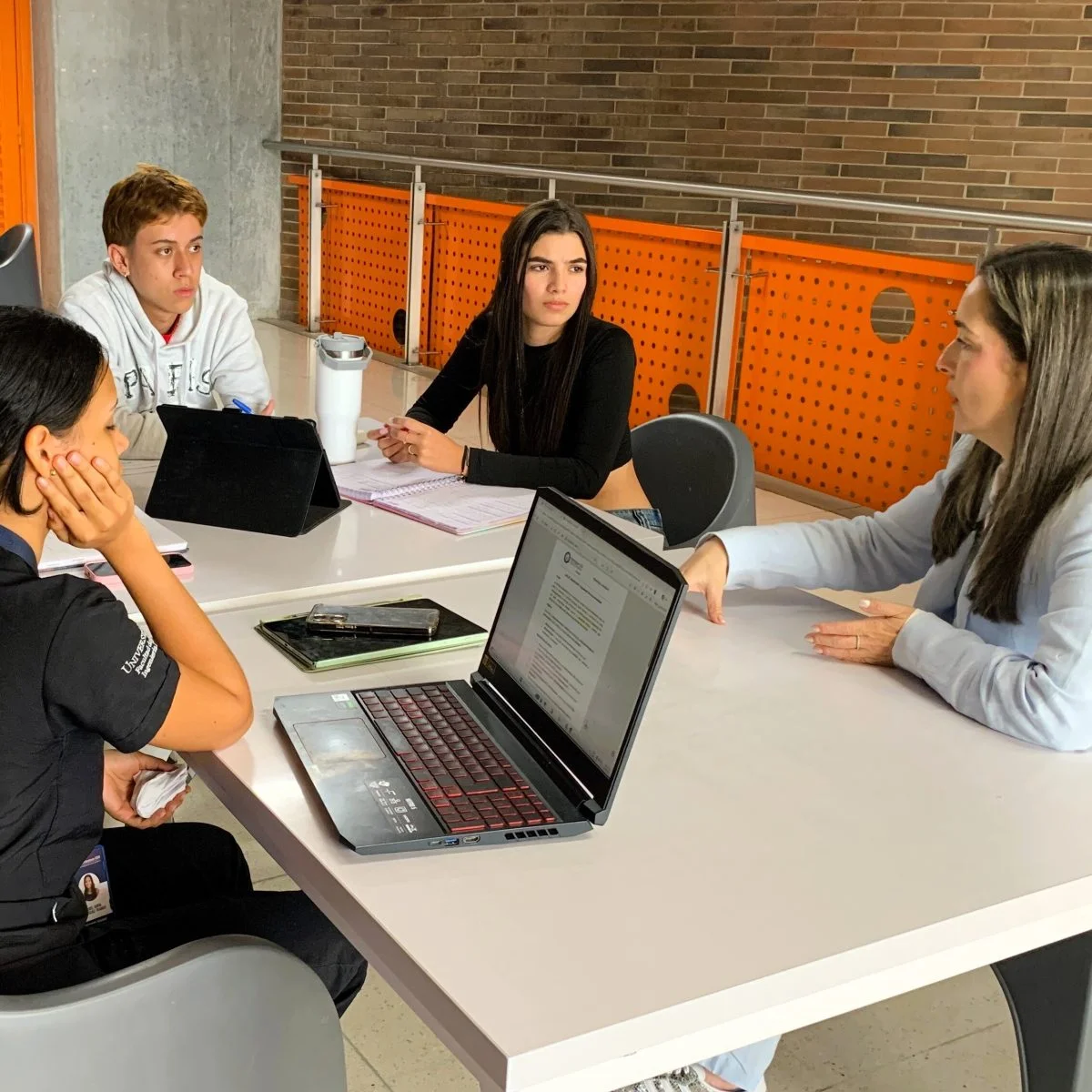 Cuatro personas participan en una reunión académica alrededor de una mesa blanca. En la mesa hay un computador portátil, una tableta, cuadernos y dispositivos móviles. Una persona realiza una intervención mientras las demás escuchan y toman apuntes en un espacio interior con paredes de ladrillo y paneles naranjas.