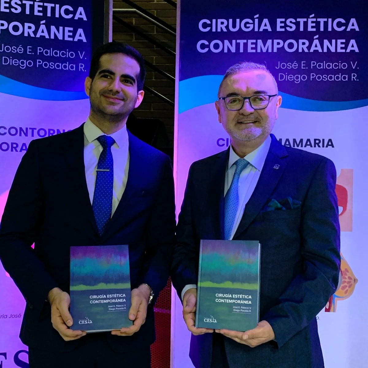 Dos personas vestidas con traje formal sostienen ejemplares del libro Cirugía Estética Contemporánea durante un evento académico. Al fondo se observan pendones promocionales con el título de la publicación y los nombres de sus autores, iluminados con luces en tonos azul y morado.