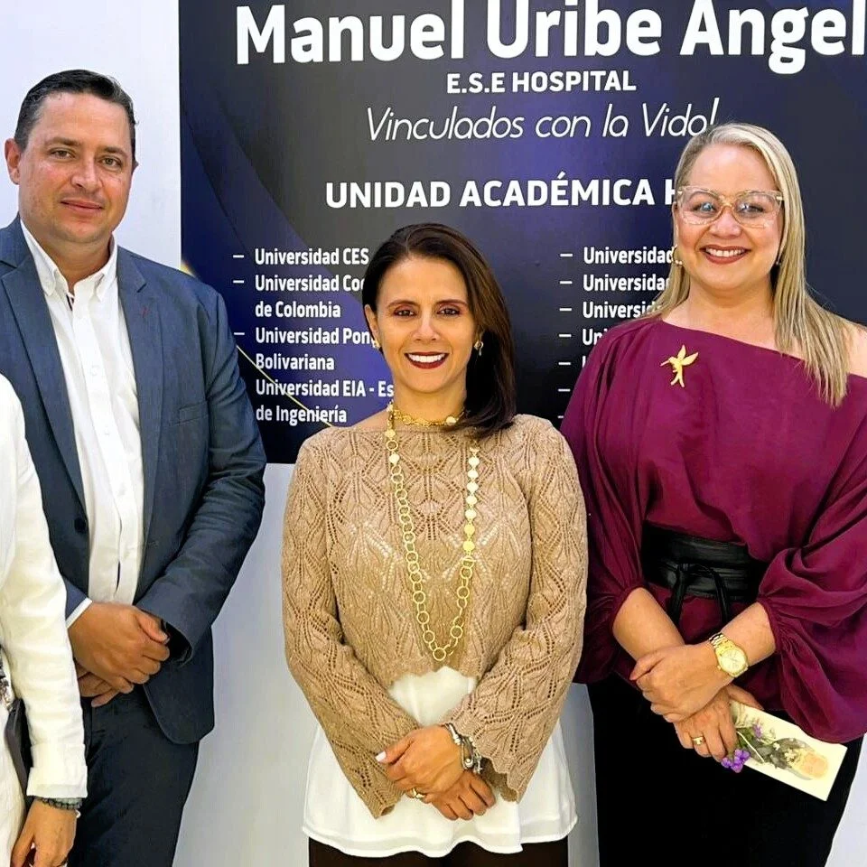 Tres personas posan de pie frente a un pendón institucional del Hospital Manuel Uribe Ángel. En el fondo se observa información sobre una unidad académica hospitalaria y los nombres de varias universidades vinculadas. Las personas visten ropa formal y están ubicadas una al lado de la otra en un espacio interior con iluminación uniforme.
