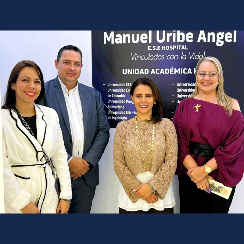 personas posan de pie frente a un pendón institucional del Hospital Manuel Uribe Ángel. En el fondo se observa información sobre una unidad académica hospitalaria y los nombres de varias universidades vinculadas. Las personas visten ropa formal y están ubicadas una al lado de la otra en un espacio interior con iluminación uniforme.