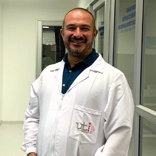 Persona de pie en un laboratorio, vistiendo una bata blanca sobre una camisa azul, junto a divisiones de vidrio y espacios de trabajo científicos en un entorno interior.