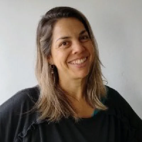Persona de pie frente a un fondo claro, vistiendo una blusa oscura de manga larga, en una fotografía tipo retrato profesional.
