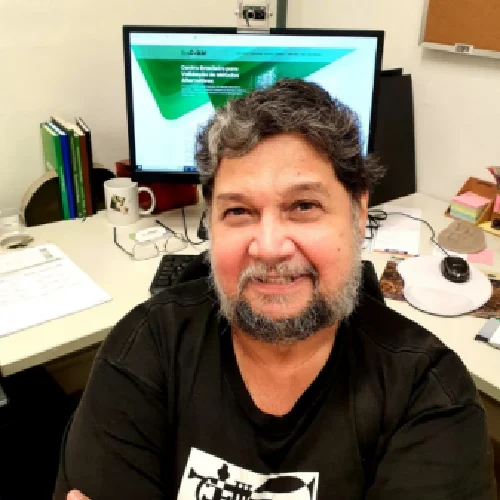 Persona sentada en un espacio de oficina frente a un escritorio con computador, documentos y materiales de trabajo, vistiendo una camiseta oscura en un entorno administrativo o académico.