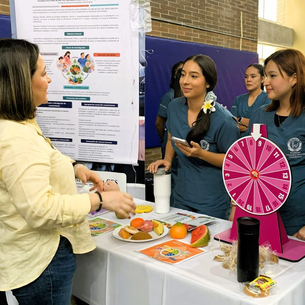 Grupo de estudiantes en uniforme participa en una actividad educativa sobre alimentación y nutrición en una feria académica. Frente a un stand informativo, observan una demostración con alimentos de muestra, material gráfico y una ruleta interactiva utilizada como recurso didáctico.