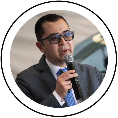 Persona en fotografía tipo retrato, centrada dentro de un marco circular, vistiendo traje gris, camisa blanca, corbata azul y gafas de montura oscura, mientras sostiene un micrófono durante una presentación en un evento.