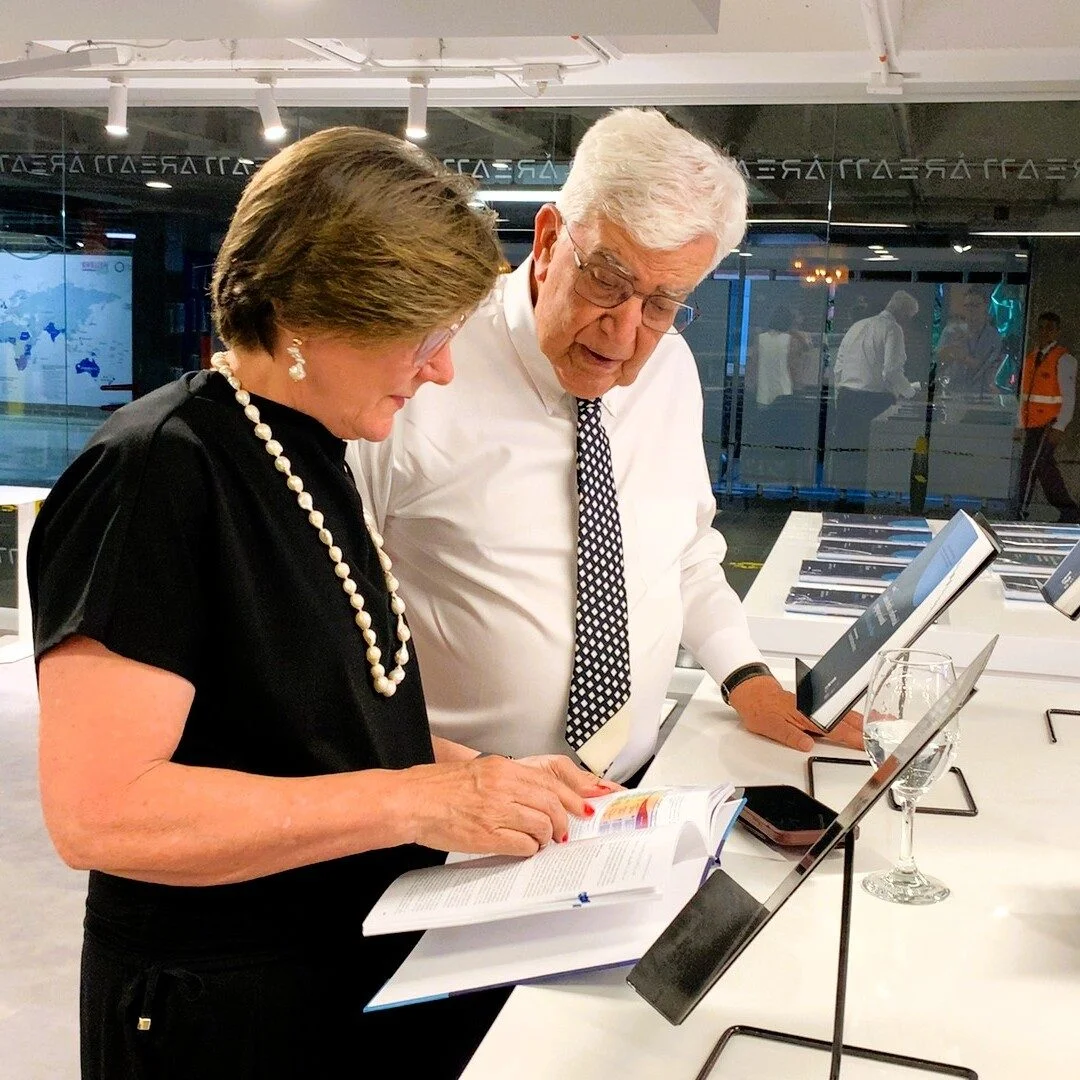 Dos personas consultan un libro abierto sobre una mesa de exhibición en un espacio interior moderno. Alrededor hay varios ejemplares colocados en soportes, iluminación superior y paredes de vidrio al fondo.