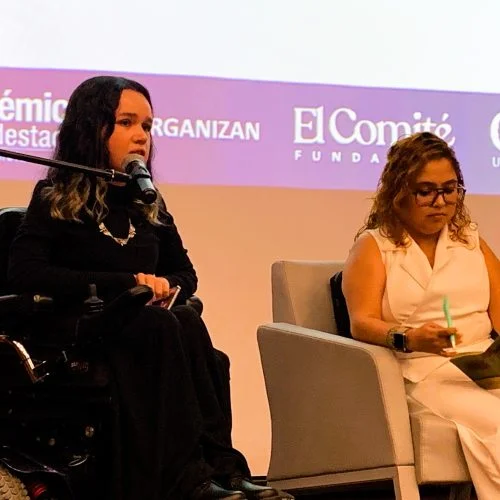 Dos personas participan en una conversación durante un evento académico. Una de ellas interviene frente a un micrófono desde una silla de ruedas motorizada, mientras la otra toma notas en una butaca; al fondo se proyectan los logotipos de instituciones organizadoras.