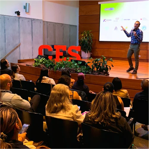 Auditorio con personas asistentes observando una presentación académica; una persona expone frente a una pantalla de proyección en un escenario con el logo ‘CES’ y elementos decorativos vegetales.