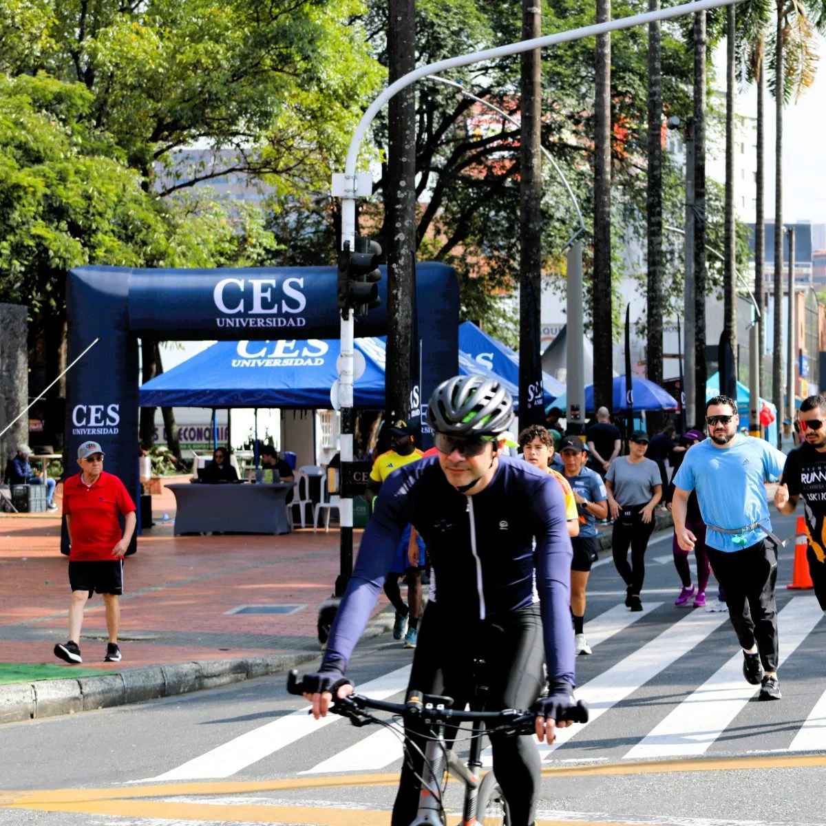 Participantes de una jornada deportiva y de bienestar realizan actividad física en una vía urbana habilitada para el evento. En primer plano, una persona se desplaza en bicicleta mientras otras caminan o corren; al fondo se observan carpas y señalización institucional de la Universidad CES.