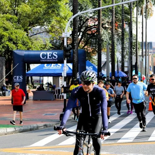Participantes de una jornada deportiva y de bienestar realizan actividad física en una vía urbana habilitada para el evento. En primer plano, una persona se desplaza en bicicleta mientras otras caminan o corren; al fondo se observan carpas y señalización institucional de la Universidad CES.