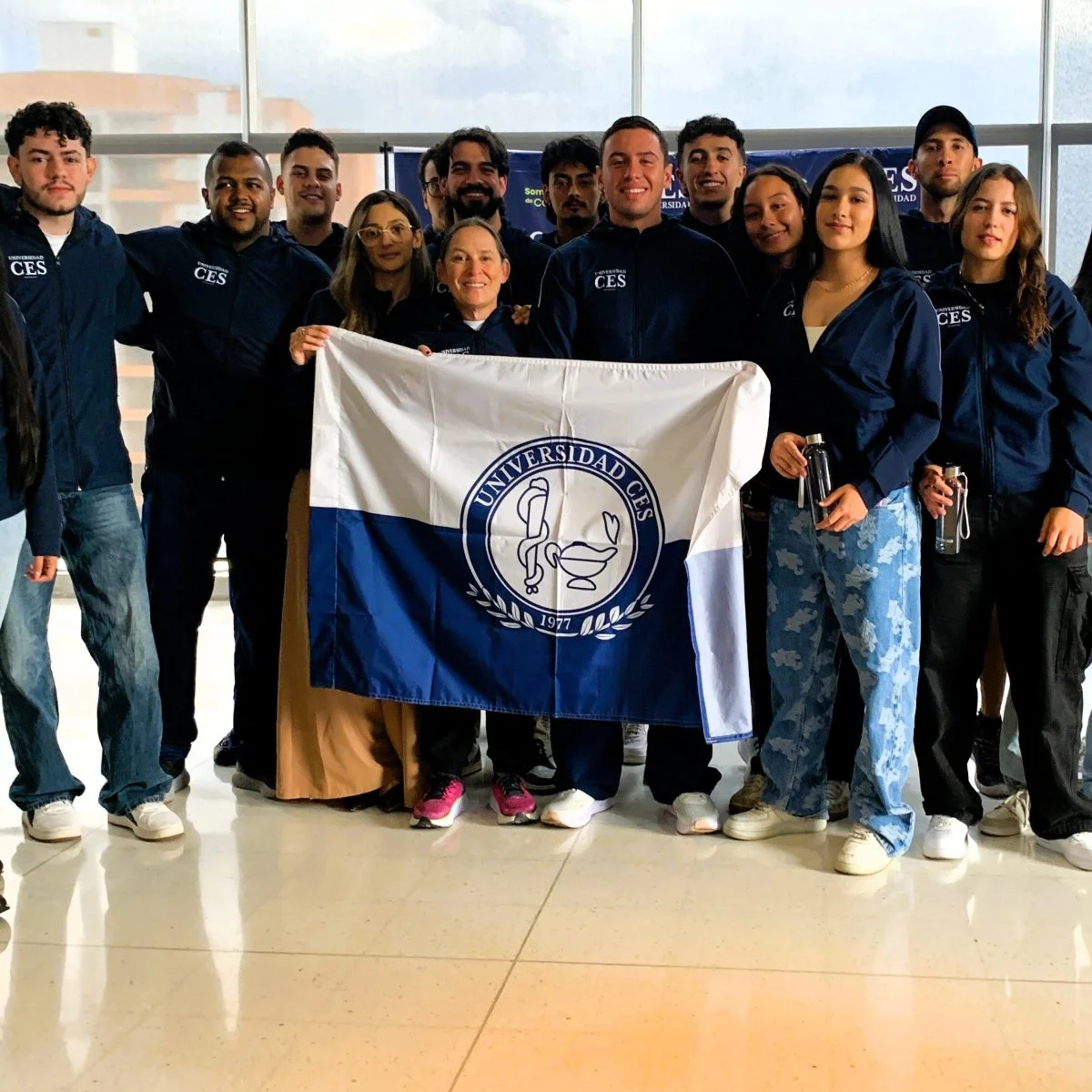 Grupo de estudiantes y docentes de la Universidad CES posan en un espacio interior sosteniendo una bandera institucional. La mayoría viste chaquetas azules con el logotipo de la universidad y se encuentran frente a ventanales que dejan ver edificaciones al exterior.