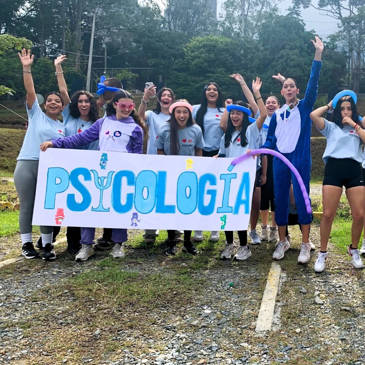 Grupo de estudiantes reunidos en un espacio al aire libre durante una actividad académica y recreativa. En primer plano sostienen una pancarta con la palabra “PSICOLOGÍA”, mientras varias personas levantan globos de colores y participan en una dinámica grupal rodeada de zonas verdes y árboles.