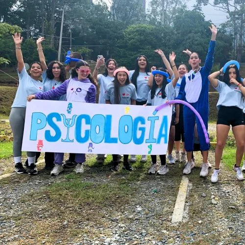 Grupo de estudiantes reunidos en un espacio al aire libre durante una actividad académica y recreativa. En primer plano sostienen una pancarta con la palabra “PSICOLOGÍA”, mientras varias personas levantan globos de colores y participan en una dinámica grupal rodeada de zonas verdes y árboles.
