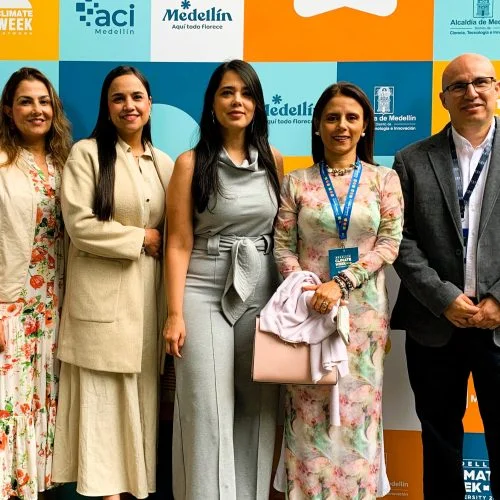Cinco participantes posan frente a un panel institucional con los logos de Climate Week, ACI Medellín y la Alcaldía de Medellín. La fotografía fue tomada en el marco de un evento académico o de innovación, en un espacio interior destinado para registro y documentación de asistentes.