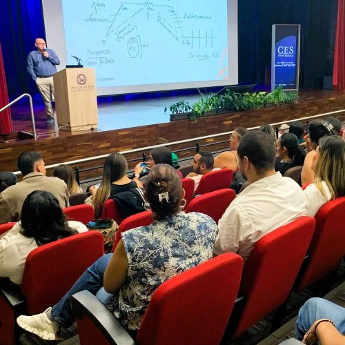Auditorio universitario con asistentes sentados en filas de sillas rojas mientras una persona realiza una presentación desde un podio. Al frente se proyecta una diapositiva en una pantalla grande durante una actividad académica.