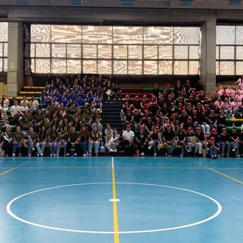 Amplio grupo de estudiantes reunidos en las graderías de un coliseo o escenario deportivo cubierto. Las personas están organizadas por equipos identificados con camisetas y accesorios de diferentes colores, en el marco de una actividad de integración o encuentro universitario.