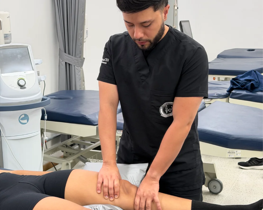 Profesional de la salud realiza una evaluación o intervención manual sobre la rodilla y la pierna de una persona recostada en una camilla, en un entorno clínico equipado para rehabilitación y atención física.