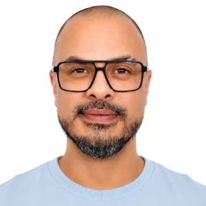Persona en fotografía tipo retrato, con gafas de montura oscura, barba corta y camiseta azul claro, sobre un fondo gris claro uniforme.