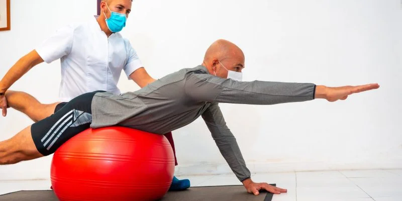 Persona realiza un ejercicio de estabilidad y fortalecimiento sobre una pelota terapéutica mientras recibe acompañamiento de un profesional de rehabilitación. La actividad se desarrolla en un espacio clínico destinado a fisioterapia y entrenamiento funcional.