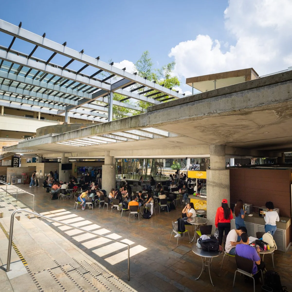 Espacio de encuentro en un campus universitario con mesas, sillas y áreas cubiertas donde estudiantes y visitantes comparten y realizan actividades. El lugar cuenta con zonas de circulación, locales de alimentos y una estructura arquitectónica abierta con iluminación natural.