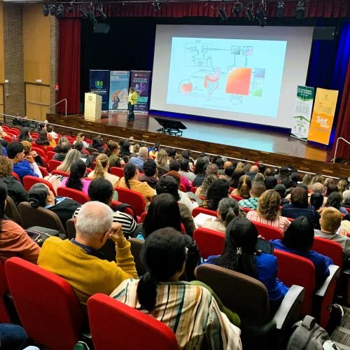 Auditorio con asistentes ubicados en sillas rojas mientras una persona realiza una conferencia en el escenario. En la pantalla principal se proyecta una diapositiva con contenido académico durante una actividad educativa.