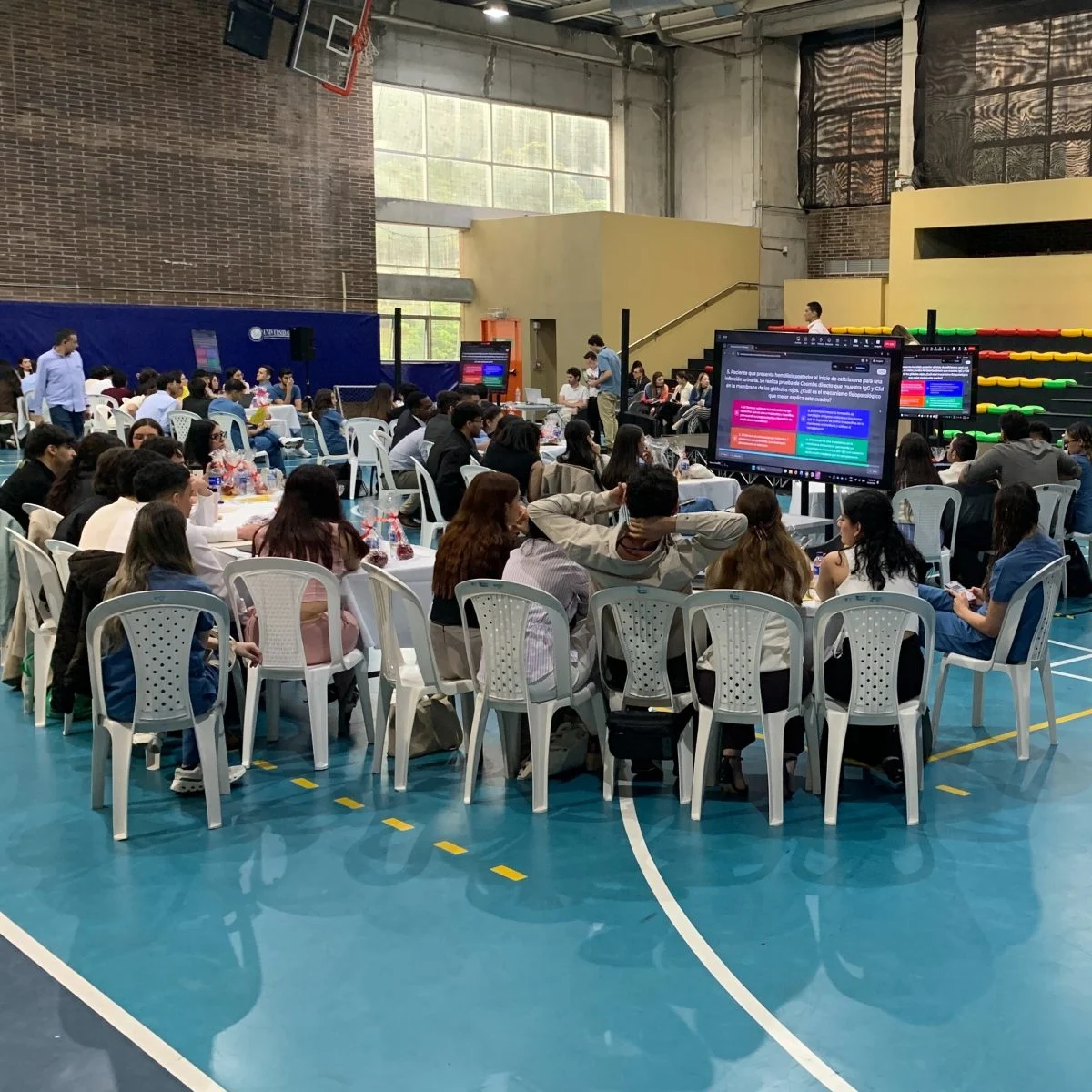 Participantes trabajan en grupos alrededor de mesas dentro de un coliseo deportivo adaptado para una actividad académica o colaborativa. Varias pantallas distribuidas en el espacio muestran información de apoyo, mientras las graderías del recinto se observan al fondo.