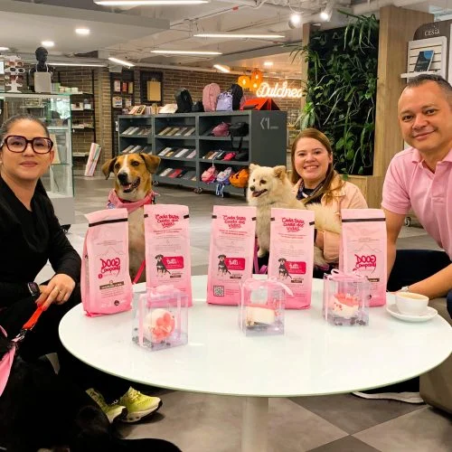 Tres personas y varios perros participan en una actividad en un espacio interior tipo tienda o centro comercial. Alrededor de una mesa se observan productos para mascotas y material promocional relacionado con una campaña de bienestar y cuidado animal.