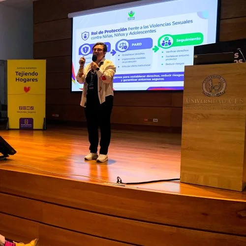 Persona expositora realiza una presentación en un auditorio de la Universidad CES ante un grupo de asistentes. En la pantalla se proyecta una diapositiva sobre protección frente a las violencias sexuales contra niñas, niños y adolescentes, junto a banderas institucionales y material informativo de una campaña social.