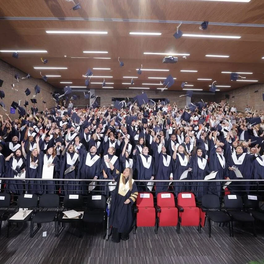 Grupo numeroso de graduandos reunidos en un auditorio durante una ceremonia de grado. Las personas visten toga y birrete y participan en el tradicional lanzamiento de los birretes al aire, mientras familiares y asistentes observan desde las graderías.