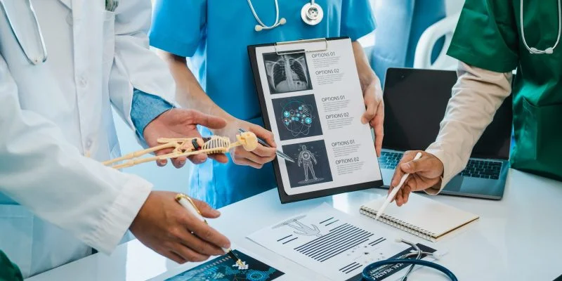 Profesionales del área de la salud revisan y analizan material clínico alrededor de una mesa de trabajo. Se observan documentos, un modelo anatómico, imágenes médicas y equipos de apoyo utilizados en procesos de aprendizaje, investigación o atención en salud.
