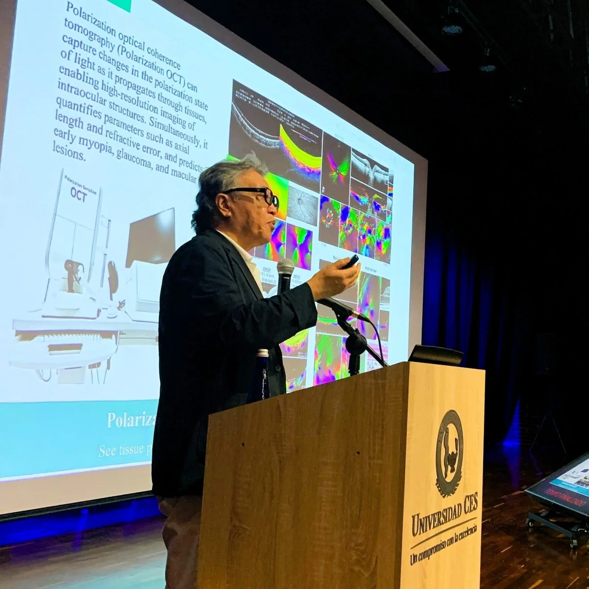 Ponente realiza una conferencia desde un atril en el auditorio de la Universidad CES. Al fondo se proyecta una presentación con imágenes y contenido relacionado con investigación y diagnóstico en ciencias de la salud, mientras los asistentes siguen la exposición.