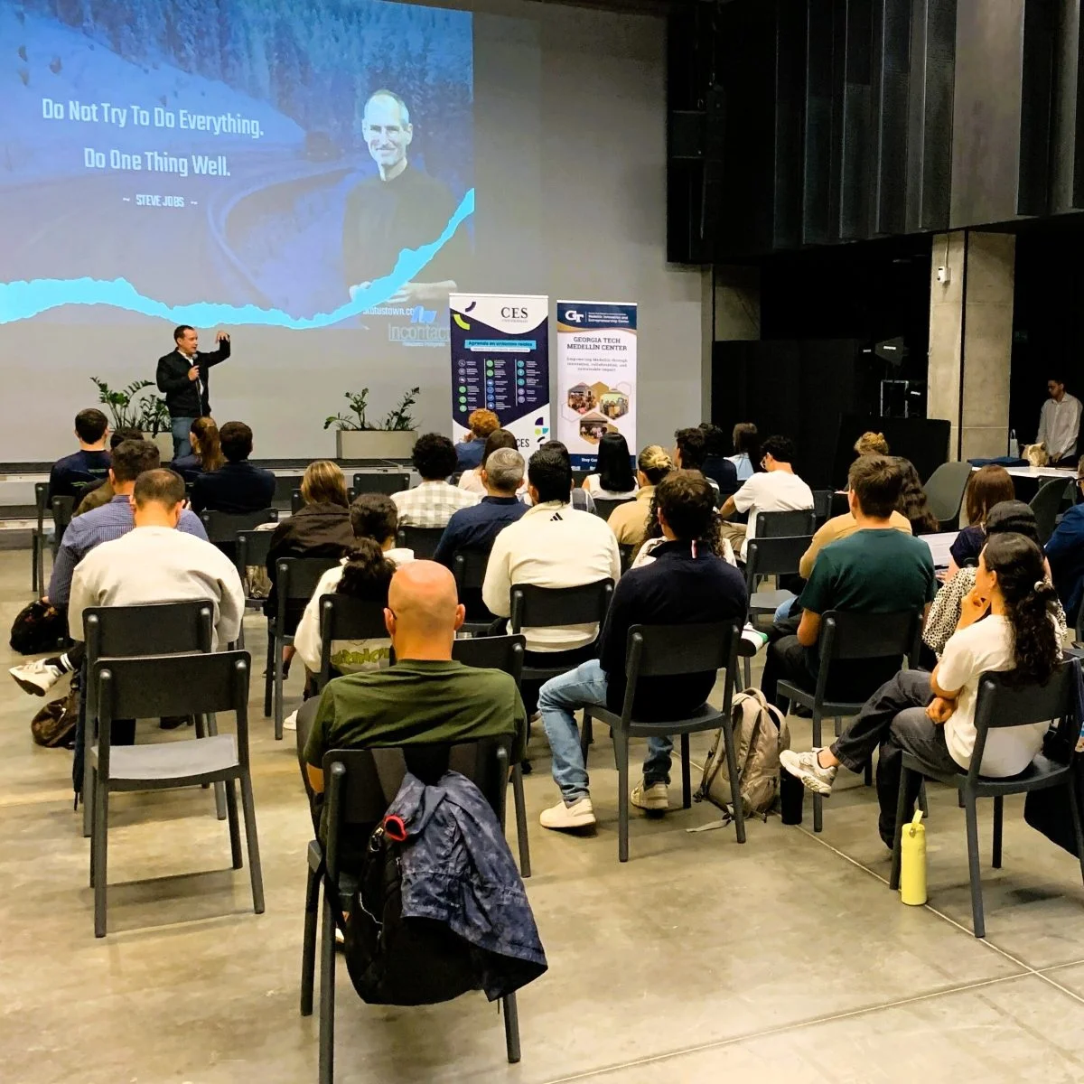 Asistentes participan en una conferencia o charla sobre emprendimiento e innovación en un auditorio. Al frente, una persona realiza una presentación mientras en la pantalla se proyecta contenido inspiracional y material relacionado con programas de la Universidad CES y aliados estratégicos.