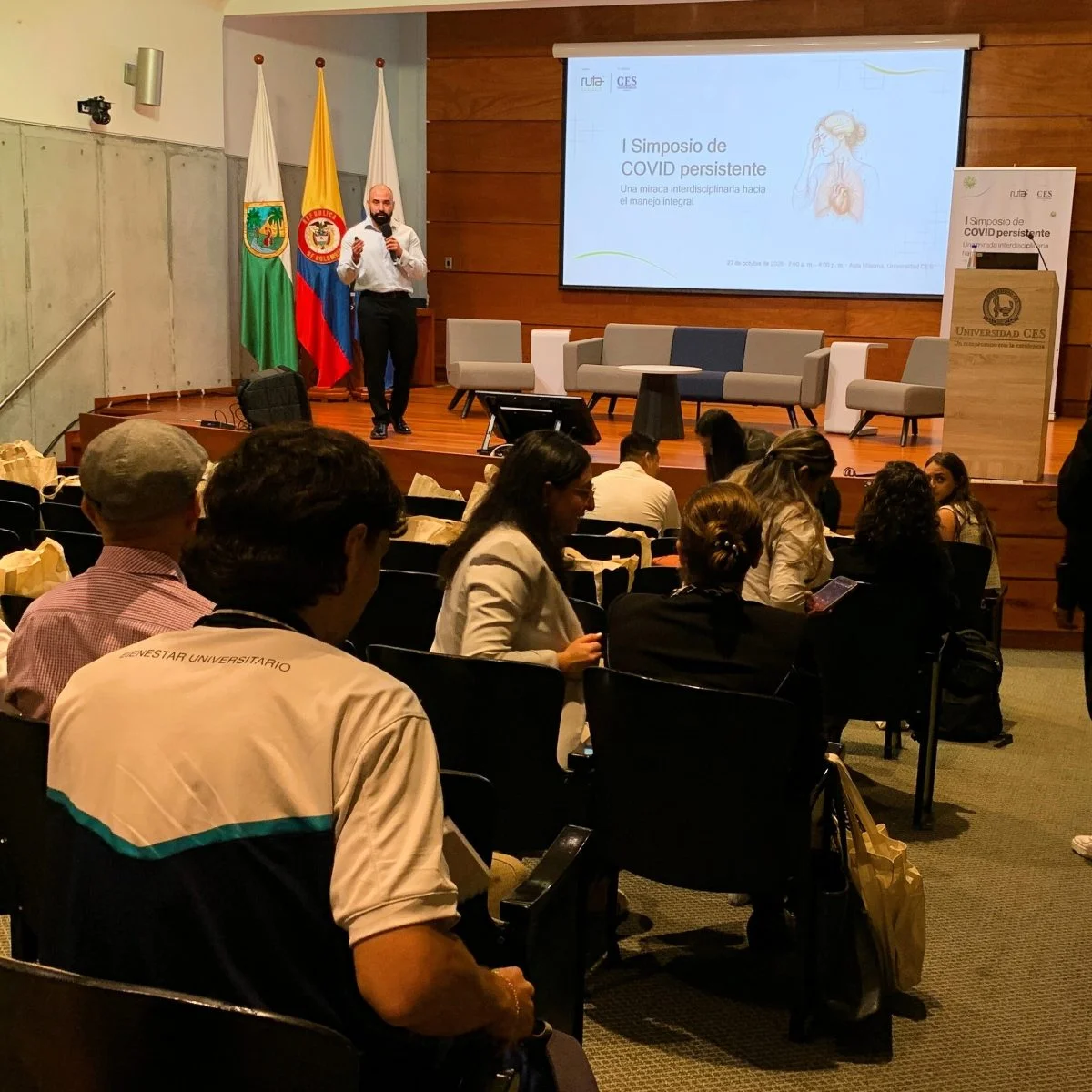 Asistentes participan en un simposio académico dentro de un auditorio mientras una persona realiza una presentación en el escenario. Al frente se proyecta una diapositiva informativa y se observan elementos propios de un evento universitario o científico.