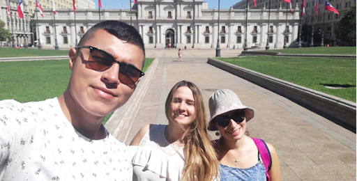 Tres personas posan para una foto en una plaza frente a un edificio gubernamental con varias banderas de Chile, en un día soleado.