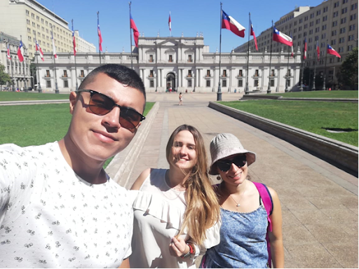 Tres personas posan para una foto en una plaza frente a un edificio gubernamental con varias banderas de Chile, en un día soleado.