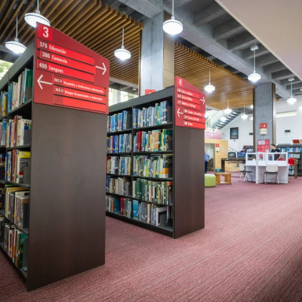 Estanterías con libros organizados en una biblioteca moderna, con señalización roja que indica categorías y direcciones, y mesas de estudio al fondo en un espacio iluminado.